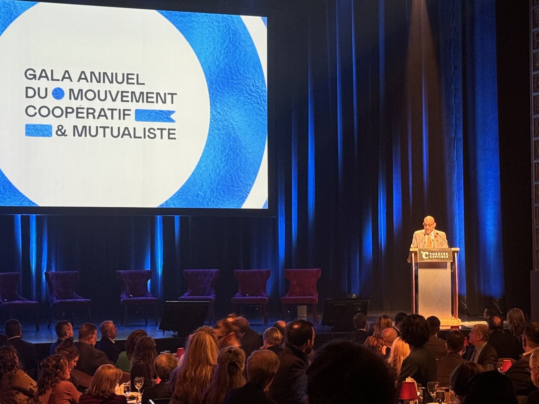 Christopher Skeete speaks at a podium before a large audience with a screen that says Gala annuel du mouvement coopératif et mutualiste.